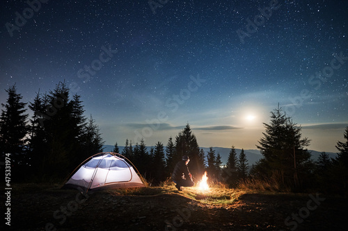Beautiful view of night starry sky over grassy hill with male hiker, illuminated camp tent and coniferous trees. Man traveler squatting down near tourist tent under night sky with stars and full moon. © anatoliy_gleb