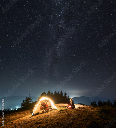 Male tourist holding guitar in hands recalsing song for his wife, being in tent in their mountain campsite. Romantic atmosphere of sky lit by stars, Milky Way and light from city under hills far away.