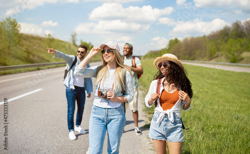 Group of millennial multiracial friends with backpacks walking along road, traveling together outdoors