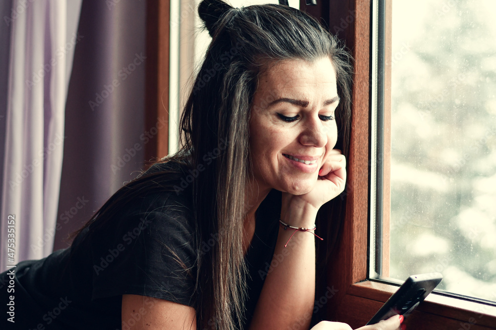 Cheerful smiling woman at home near the window at winter time looking at the phone reading a Message ,checking social media,playing game, shopping online Christmas gifts ordering delivery Cheerful smiling woman at home near the window at winter time looking at the phone reading a Message ,checking social media,playing game, shopping online Christmas gifts ordering delivery
