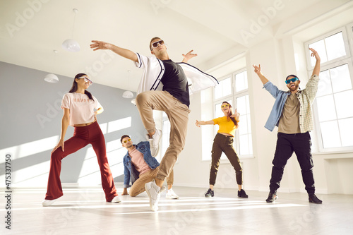 Group of people dancing in modern studio. Energetic male dancer with cool, free attitude, wearing trendy outfit, mixing styles and doing ballet fouette during break dance class with friends, low angle