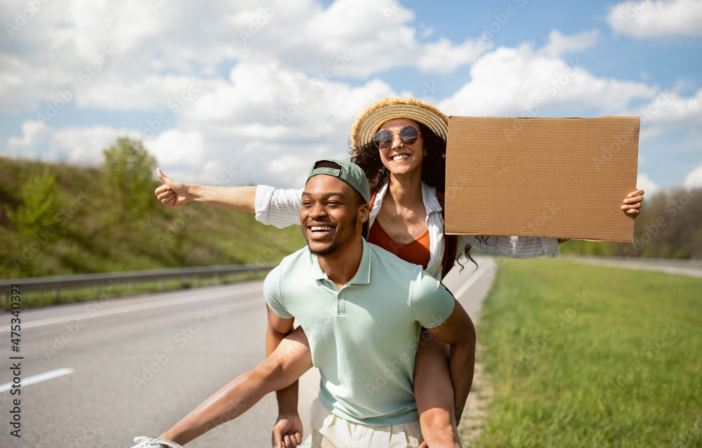 Obraz premium Happy young multiracial couple hitchhiking together, holding empty cardboard sign, showing thumb up gesture outdoors