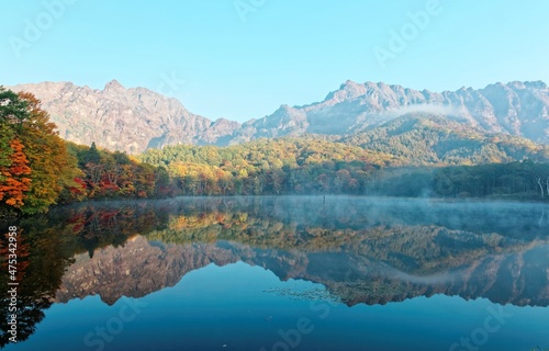 Autumn scenery of amazing Kagami Ike (Mirror Pond) in morning light with symmetric reflections of colorful fall foliage on smooth lake water & rugged Togakushi Mountain in background in Nagano, Japan