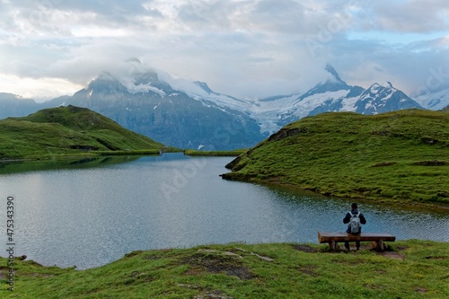 Tourist sitting on a bench by the grassy lakeside of Bachalpsee & enjoying the panoramic view of majestic alpine mountains under moody cloudy sky in First, Grindelwald, Bernese Highlands, Switzerland