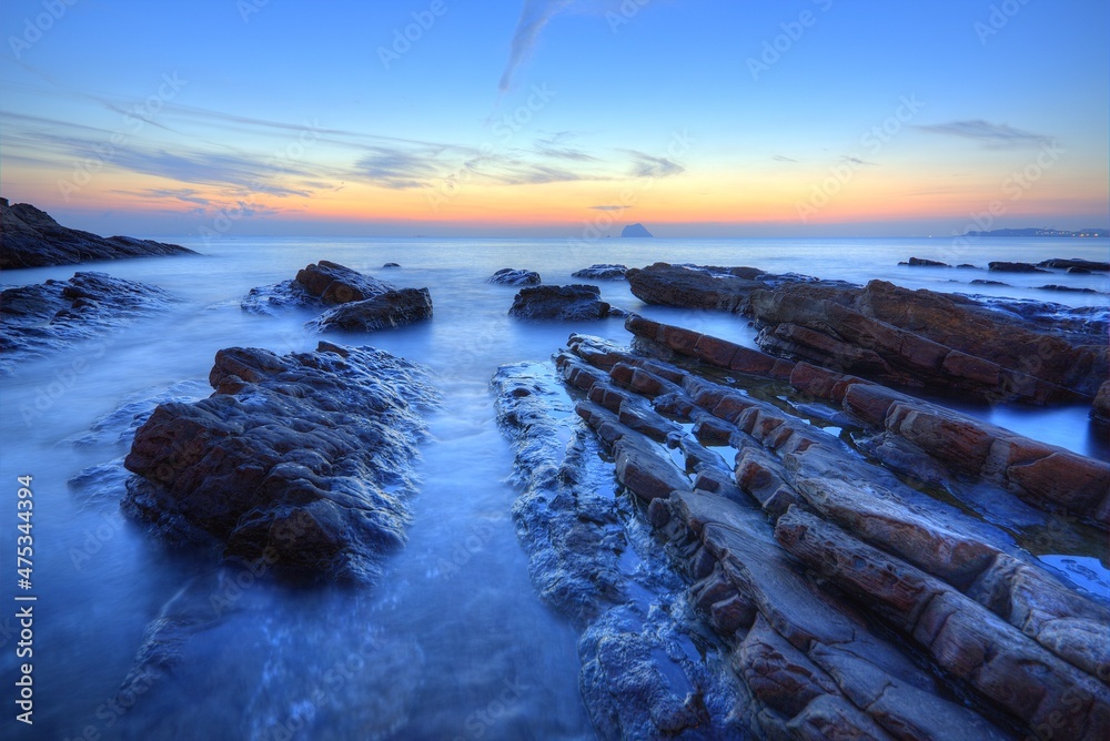 Obraz premium Beautiful sunrise scenery of a rocky beach on northern coast of Taiwan with an island on distant horizon & peculiar rock formations on seashore under dramatic dawning sky (Long Exposure Effect)