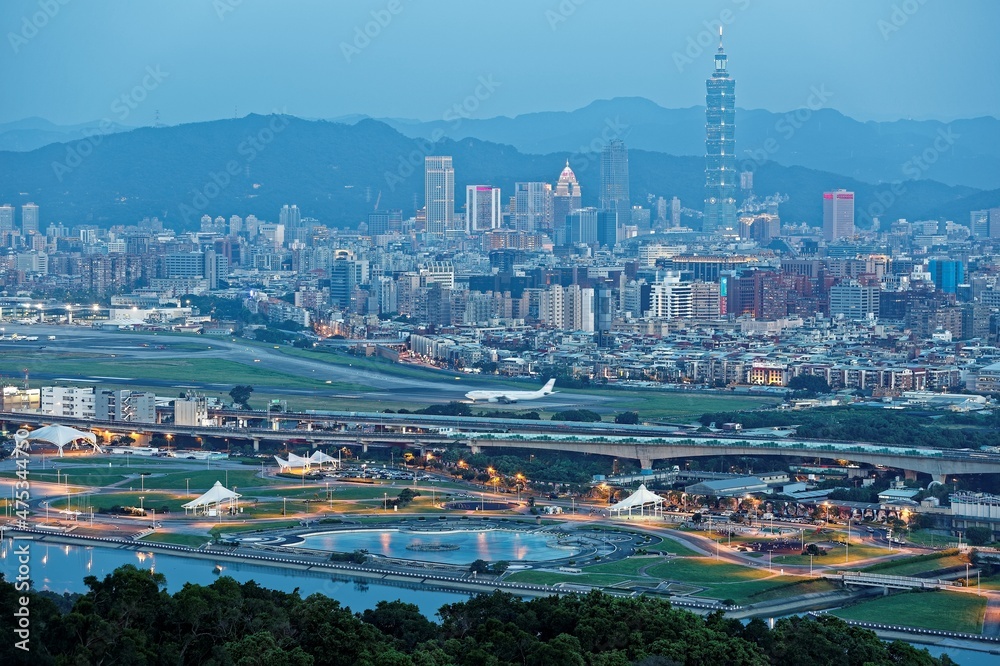 Aerial panorama of Taipei in evening twilight, capital city of Taiwan, with Taipei 101 Tower amid skyscrapers in downtown, a riverside park by Keelung River & an airplane parking in Songshan Airport