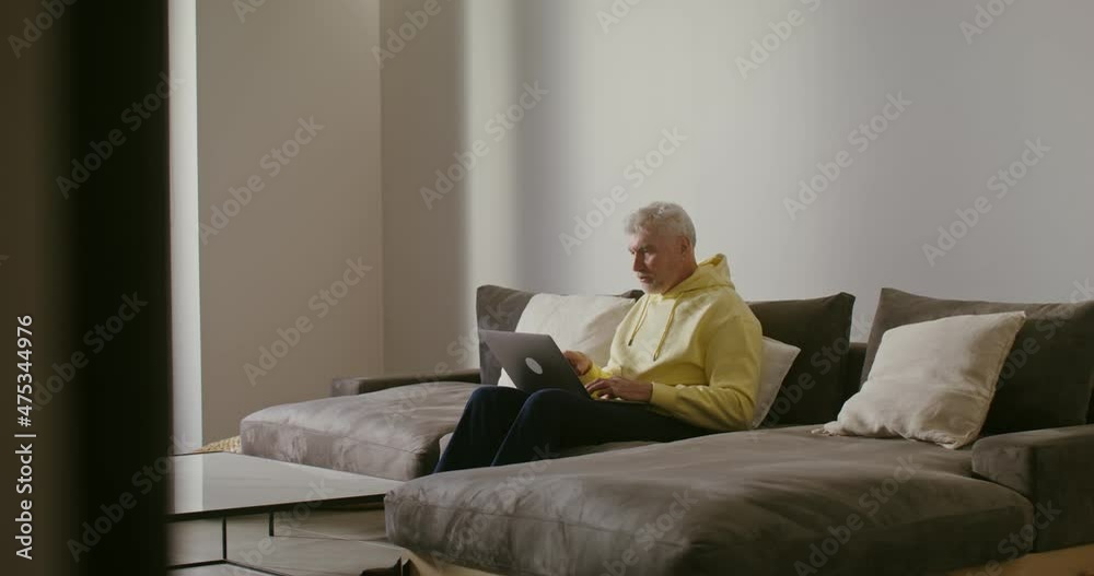 An elderly man smiling starts video call, waving hello sitting on a sofa at home An elderly man smiling starts video call, waving hello sitting on a sofa at home