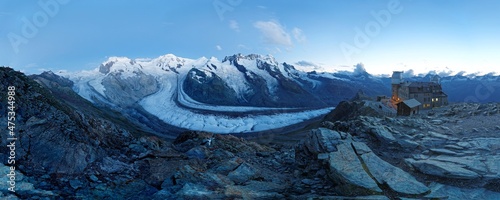 A panorama of Gorner Glacier with the moon shining over snow capped mountains (Monte Rosa Massif, Liskamm & Matterhorn) and the Gornergrat Observatory on a rocky ridge in Zermatt, Valais, Switzerland photo