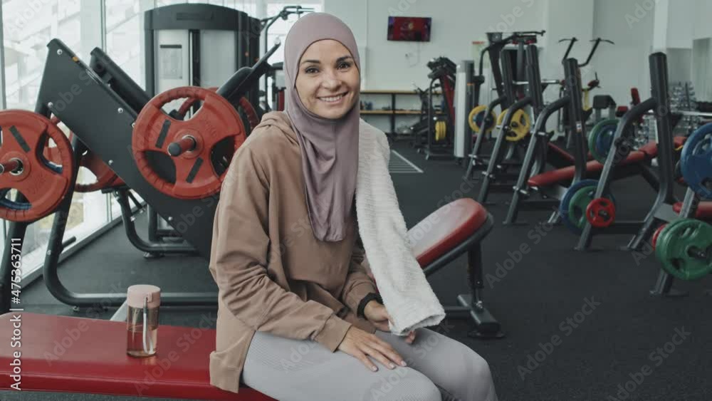 Portrait of beautiful Muslim woman in hijab wiping face with towel, then looking at camera and smiling while resting in gym after workout