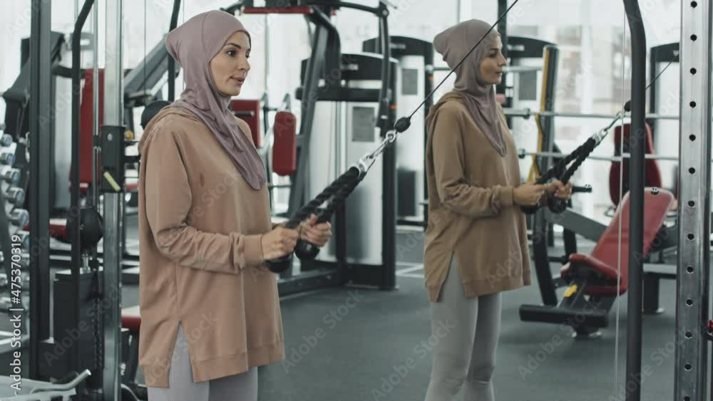 Muslim woman in hijab and sportswear doing standing pull downs with cable machine and looking in gym mirror