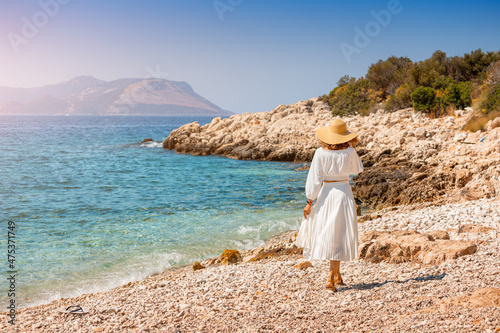 Happy and elegant woman traveler in hat and a charming white dress is walking on the sea shore of a tropical resort. Vacation and tourism concept. photo