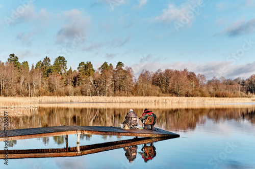 Two fisherman with fishing rods sitting on wooden bridge among scenic lake, reflecting on mirror surface of water.