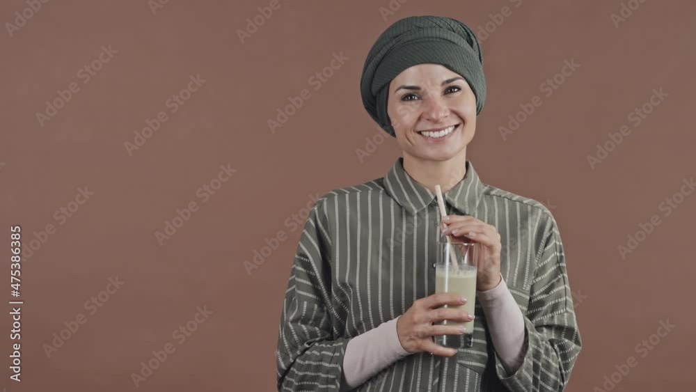 Portrait of beautiful Muslim woman in hijab drinking fresh smoothie and smiling at camera while posing in studio