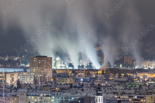 Night city view with buildings and a smoking industrial steel mill with impressive exhaust gases