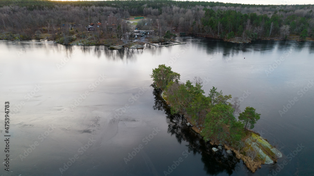 Beautiful Scandinavian landscape photographed with a drone on a sunny late autumn or winter day. Island with evergreen trees in the middle of frozen lake. Beautiful Scandinavian landscape photographed with a drone on a sunny late autumn or winter day. Island with evergreen trees in the middle of frozen lake.