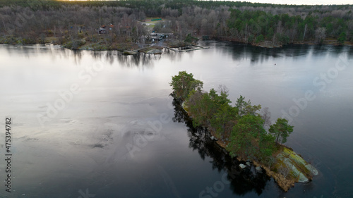 Beautiful Scandinavian landscape photographed with a drone on a sunny late autumn or winter day. Island with evergreen trees in the middle of frozen lake.