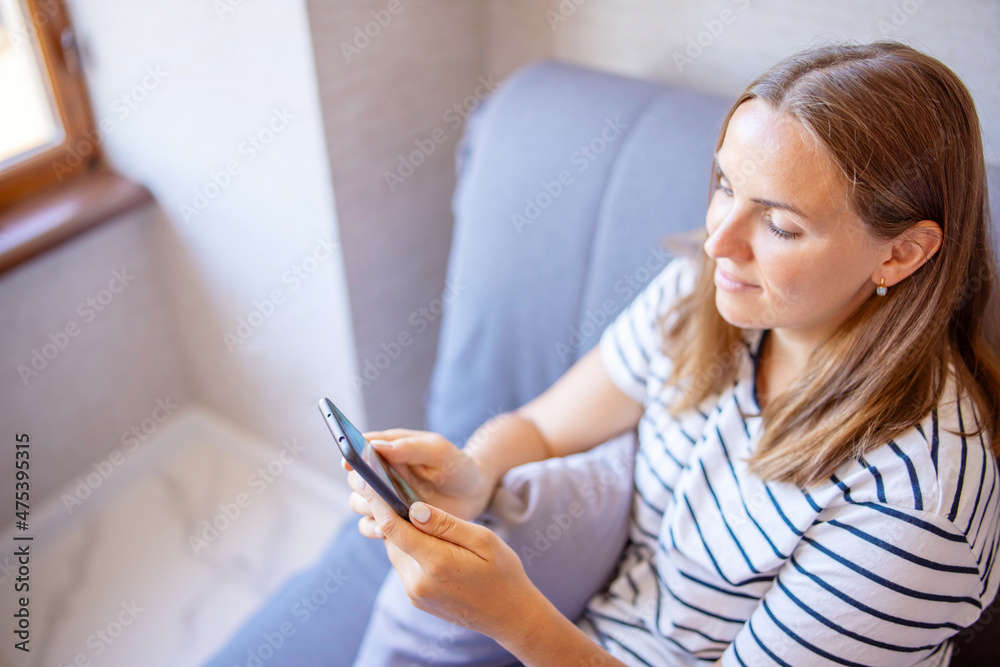 Happy young woman is using mobile phone while sitting a couch at home. A woman on couch at home message text on modern cellphone, shopping online via website Happy young woman is using mobile phone while sitting a couch at home. A woman on couch at home message text on modern cellphone, shopping online via website