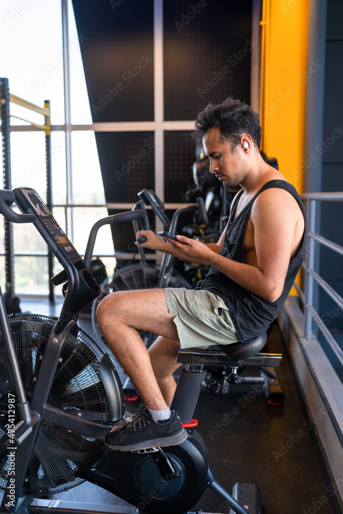 Young man cheking his phone while he is on a cycling machine in the gym Young man cheking his phone while he is on a cycling machine in the gym