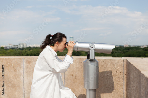 Caucasian female standing on building rooftop looking through telescope at metropolitan urban city admiring beautiful place. Panoramic landscape view from tower observation point
