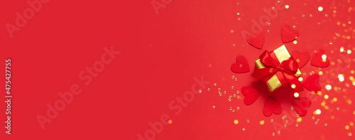 Gift with a red bow on a red background with glitter hearts and confetti. Festive flat composition for Valentine's Day, New Year, Mother's Day, Wedding. Mockup with copy space