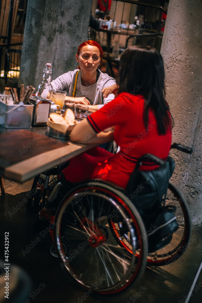 Two girls in wheelchairs in cozy cafe waiting for order. Accessible common areas for people with special needs. Affordable tourism for people with special needs due to physical special needs