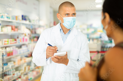 Male pharmacist in face mask writing recipe for woman while they standing in salesroom of drugstore.