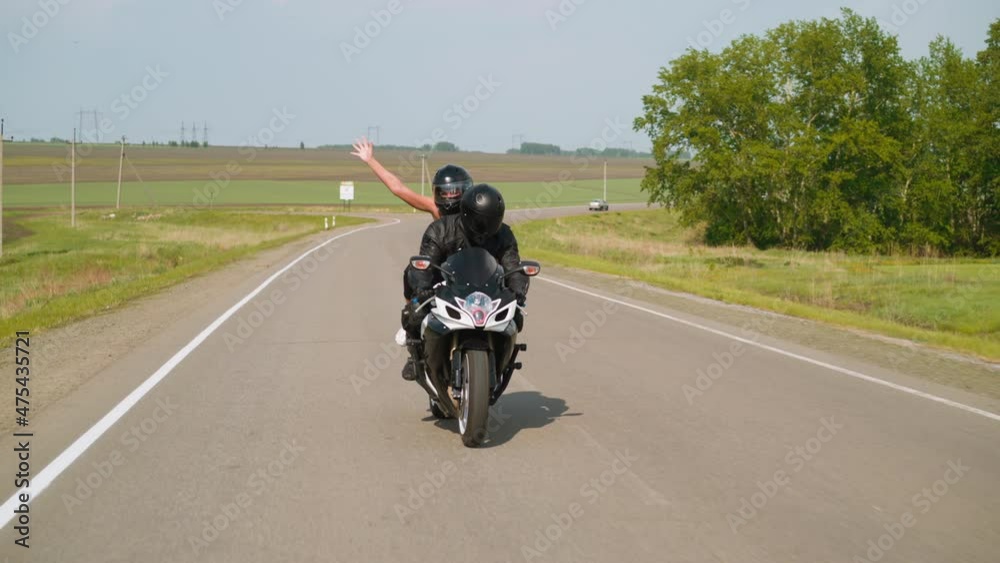 Joyful woman with raised arm enjoys motorcycle riding with boyfriend along road past green trees and empty field at countryside slow motion Joyful woman with raised arm enjoys motorcycle riding with boyfriend along road past green trees and empty field at countryside slow motion