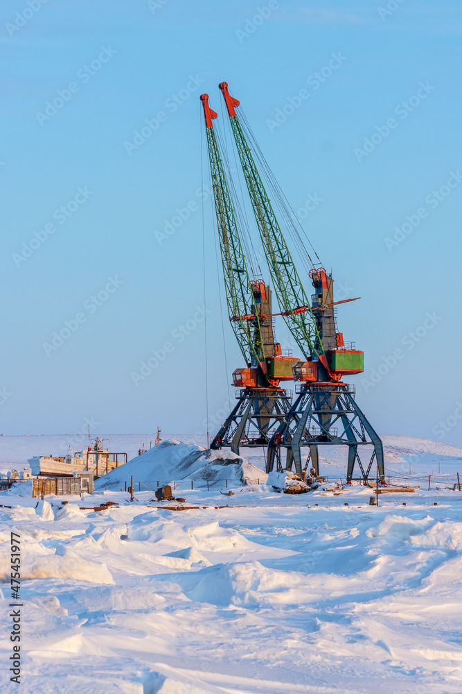 View of the port cranes on the pier among the ice and snow. Winter industrial landscape. Transport infrastructure in Siberia and in the Far North of Russia in the Arctic. Anadyr, Chukotka, Russia.