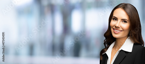 Portrait of happy smiling young businesswoman in black confident suit, over blurred modern office interior background. Business woman indoors. Copy space blank area for text.