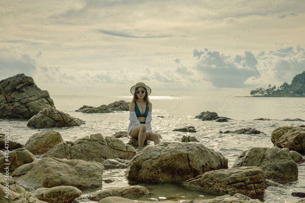 A beautiful Asian woman in a bikini walks amongst the beach rocks where the golden glow of the rising sun.