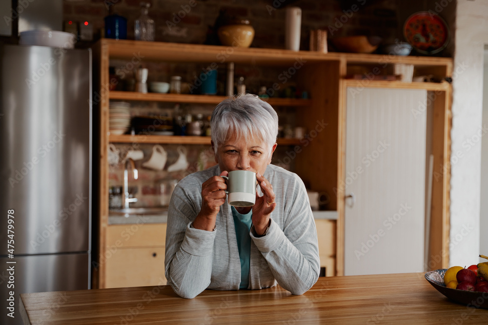 Portrait of retired biracial elderly female leaning on modern kitchen counter smiling at camera, taking sip of morning coffee.
