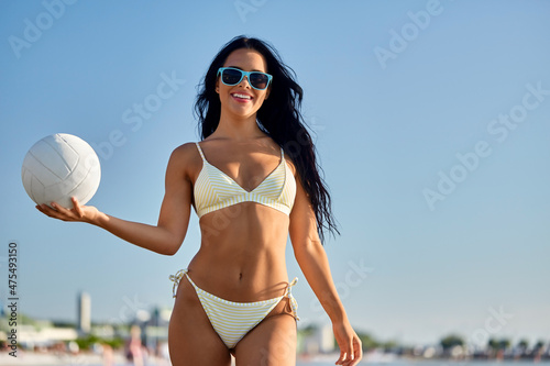 people, summer and leisure concept - happy smiling young woman in bikini swimsuit posing with volleyball on beach