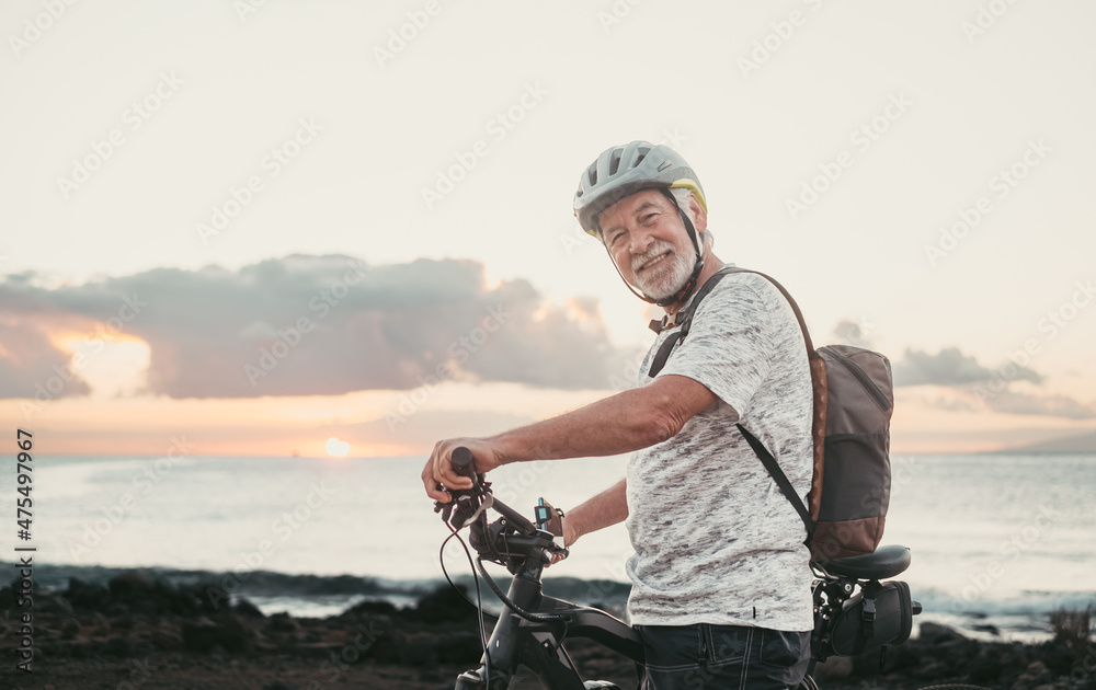 Portrait of senior cyclist man at the beach at sunset enjoying freedom and beauty in nature. Elderly man with protective helmet and backpack looking at camera Portrait of senior cyclist man at the beach at sunset enjoying freedom and beauty in nature. Elderly man with protective helmet and backpack looking at camera