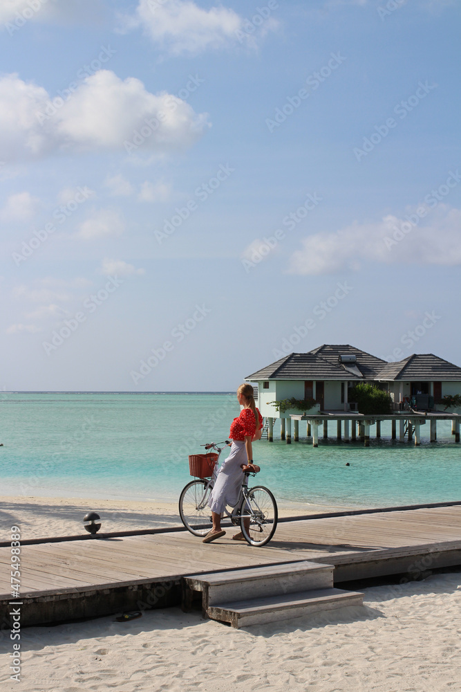 Fototapeta premium A girl in a red blouse and a white skirt stands with a bicycle on a wooden pier leading to villas on stilts above the water in the Maldives.