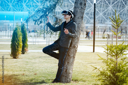 Young  man dressed in virtual reality goggles in a city park near green young spruces and trees, leaning against a tree and manipulating his hands, from behind strongly unwinding background
