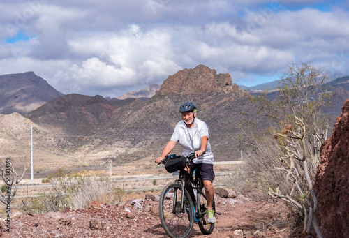 Happy senior man riding in a country road with his bike, cyclist wearing sport helmet. Mountains in background