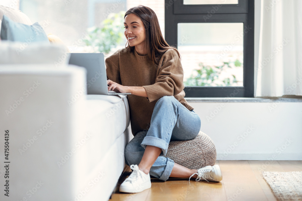 Pretty young woman working with her laptop on couch while sitting on floor in the living room at home. Pretty young woman working with her laptop on couch while sitting on floor in the living room at home.
