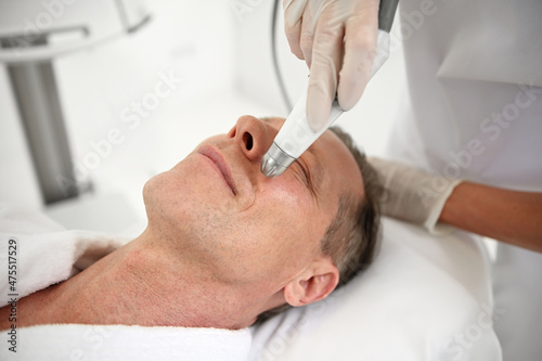 Rejuvenating facial treatment. Mature man getting lifting therapy massage in a beauty SPA salon. Exfoliation, stimulation and hydration. Men's aesthetic cosmetology, closeup, copy ad space
