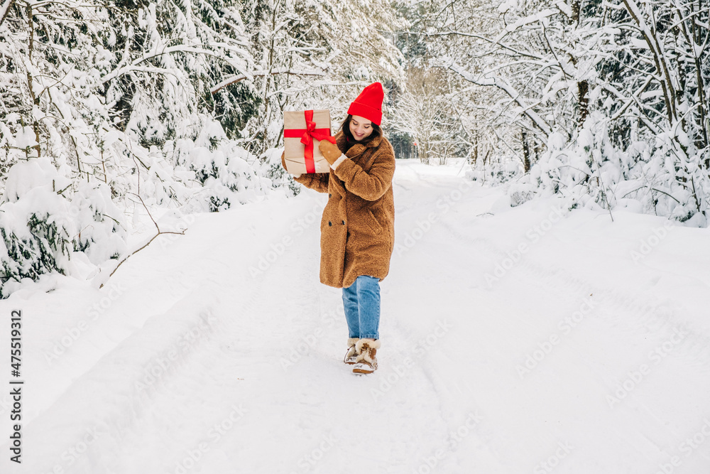 Winter Christmas shopping concept. Woman holds a large gift box with a red ribbon and walks forward in snow winter forest. 