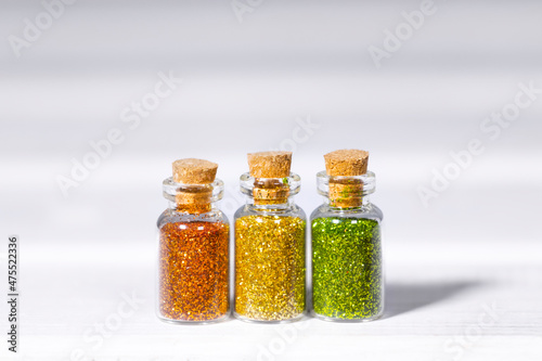 Small Glass jars with golden, orange and green glitters for nail art and makeup on white background, Copyspace