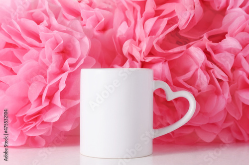 White mug on a pink paper flowers background with heart shaped handle for Valentine's day or Mothers day, template for your design.