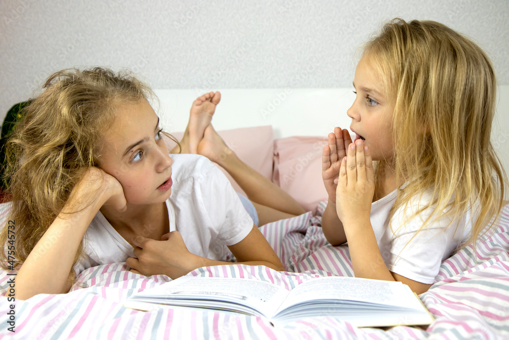 two cute little sisters with blond hair lie on the bed and read a book, tell scary stories and scare them with stories