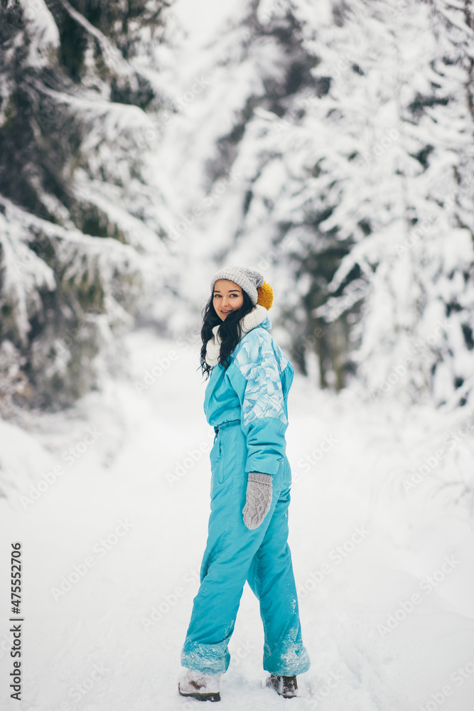 Young woman winter portrait in full height outdoors. Adorable young adult girl in ski suit walking in the mountain snowy forest.