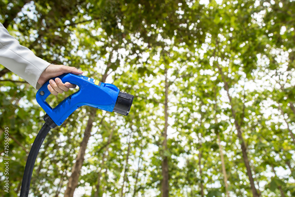 Naklejka premium Hand holding Electric car charger. Electric Vehicle EV Charging station on blurred nature with soft light background. Air pollution and reduce greenhouse gas emissions concept. Eco-friendly energy.