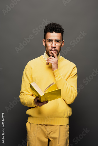 pensive african american man in glasses and yellow outfit holding book on dark grey.