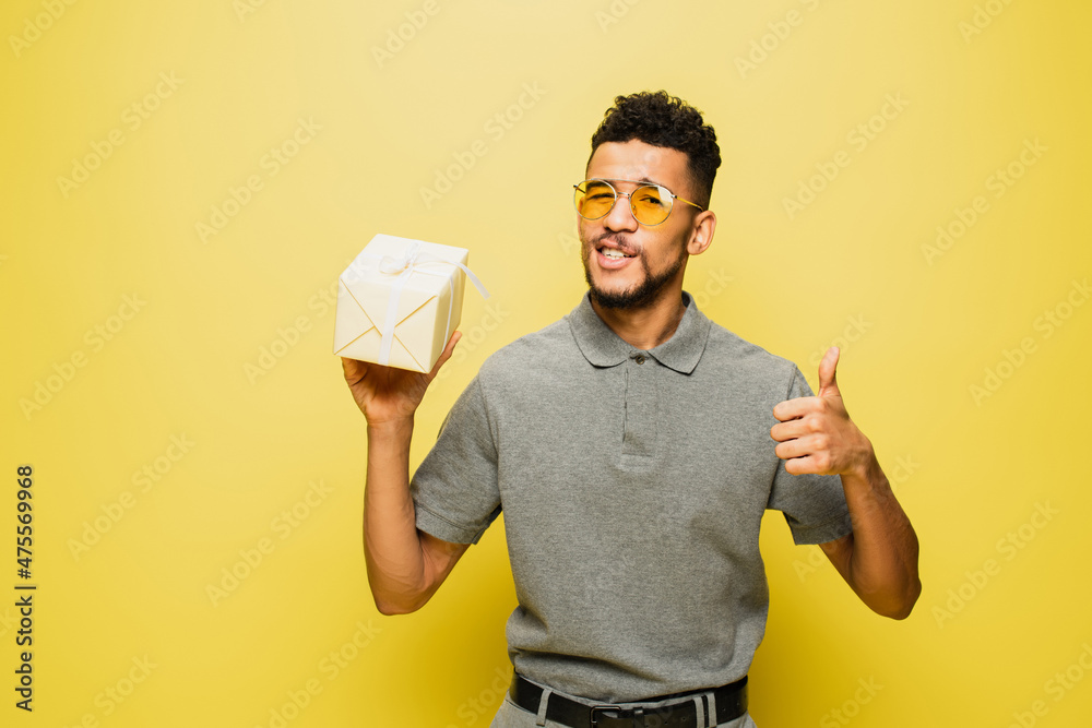 african american man in sunglasses and grey tennis shirt holding wrapped present and showing thumb up on yellow. african american man in sunglasses and grey tennis shirt holding wrapped present and showing thumb up on yellow.