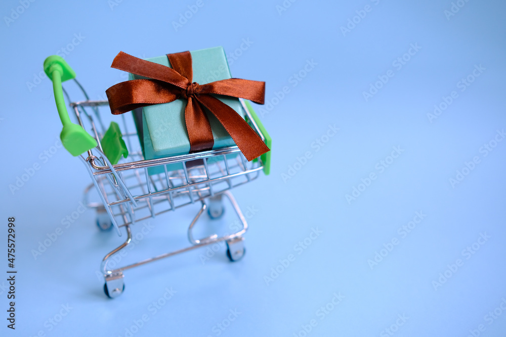 A present box with a ribbon in a shopping trolley cart on blue background . Concept - buying a gift ,online and sale. Christmas, valentines ,women day .Copy space, mockup.