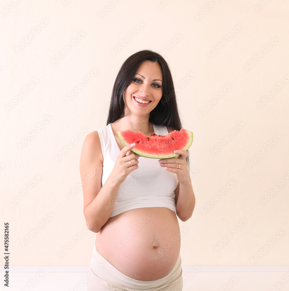 Beautiful young pregnant brunette woman with watermelon on light beige wall background with copy space. Beautiful young pregnant brunette woman with watermelon on light beige wall background with copy space.