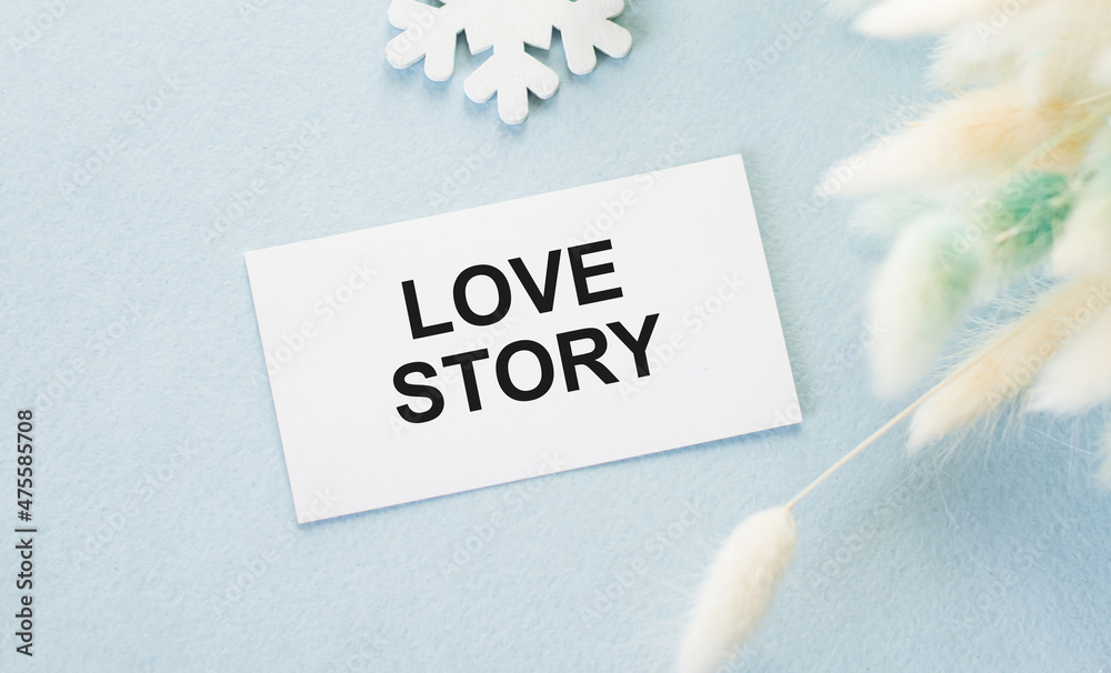 Love Story inscription on a card that lends on a blue background next to fluffy branches of dried flower, festive background Love Story inscription on a card that lends on a blue background next to fluffy branches of dried flower, festive background
