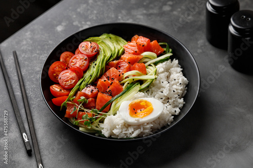Salmon Poke bowl with avocado, cherry, egg and rice on dark background with chopsticks.